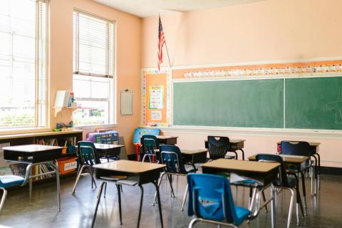 Empty elementary school classroom with green chalkboard, American flag, windows, bins on the side, and desks 