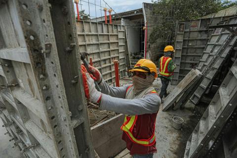 Two construction workers wearing safety gear, including hard hats and reflective vests, work with metal formwork panels at a building site.